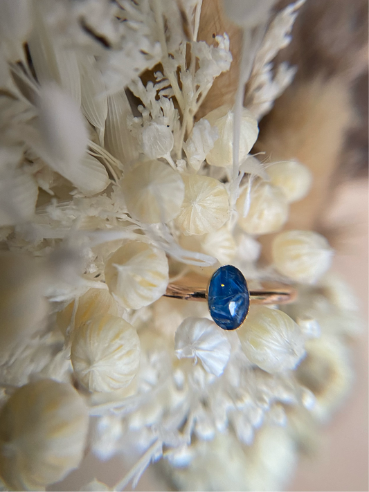 A close-up image of a small oval-shaped ring with a blue stone, surrounded by gold-filled or recycled sterling silver material, set against a floral background.