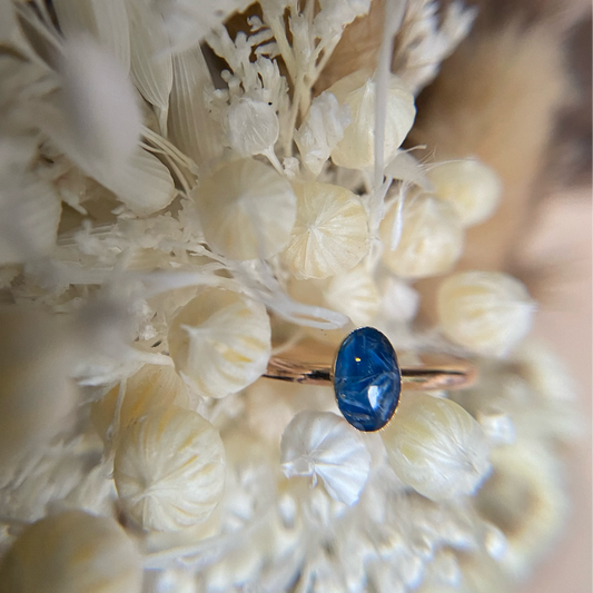 A close-up image of a small oval-shaped ring with a blue stone, surrounded by gold-filled or recycled sterling silver material, set against a floral background.
